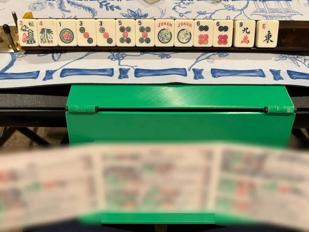 An open Mahjong card spread across the ledge of a green card holder hanging from a game table, below a rack of tiles